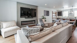 Una sala moderna con un televisor grande, chimenea y comedor, con una cocina al fondo.