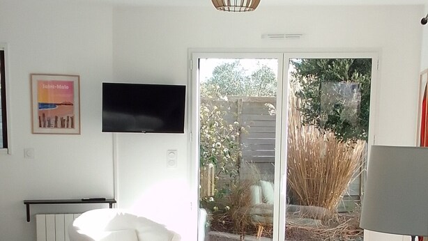 A modern living room with a white sofa, a dining table, and a TV mounted on the wall. - Maison à 50 m de la mer (Saint-Jacut-de-la-Mer)