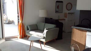 A compact living space with a sofa, a small table, a kitchen area with a dishwasher, and a view through an open door. - Maison à 50 m de la mer (Saint-Jacut-de-la-Mer)