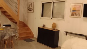 A dining area with a round table, chairs, a lamp, a staircase, a cabinet, and a window with blinds. - Maison à 50 m de la mer (Saint-Jacut-de-la-Mer)