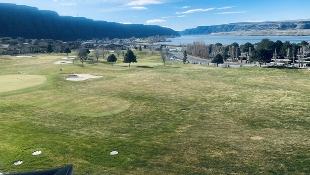 Golfkenttä, jossa on hiekkabunkkereita, puita ja näkymä järvelle ja vuorille.