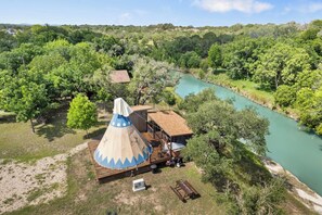 A cabin with a conical roof structure, surrounded by a river and dense greenery. - Stronghold Tipi | Creek Access & Hot Tub & Fishing (Bandera)