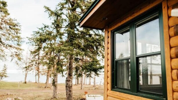 A log cabin with a large window, surrounded by trees and a grassy area. - Escape to Tranquility at The Sandy Pearl: A Cozy Log Cottage Retreat (Tatamagouche)