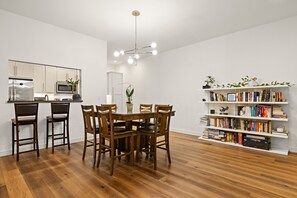 Una cocina moderna con armarios blancos, un comedor con mesa y sillas de madera, y una estantería llena de libros y objetos decorativos.