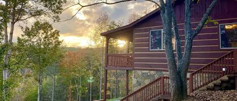 A cabin with a stone foundation, a wooden deck, and a staircase leading to a second-story balcony.