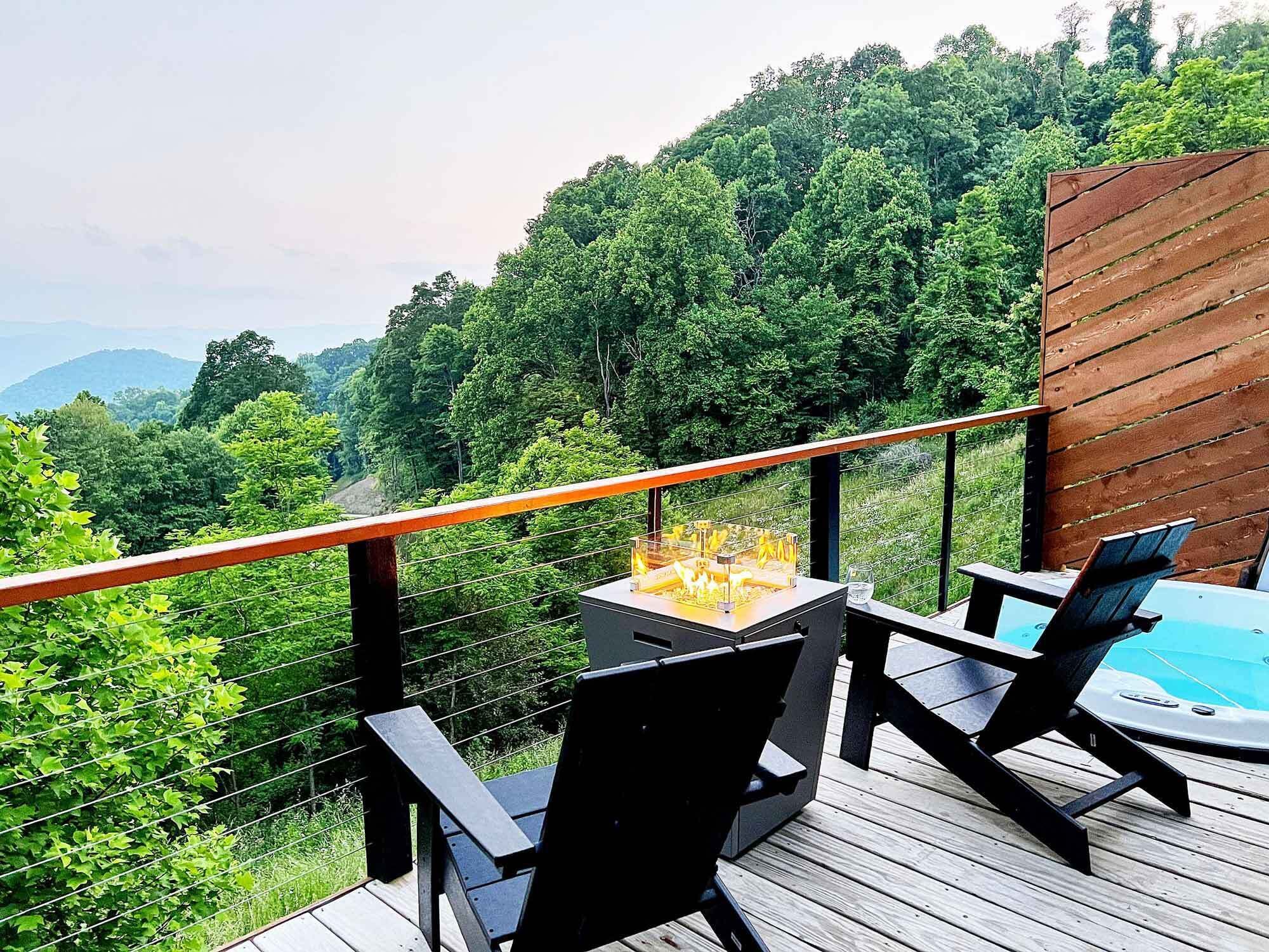 A deck with a hot tub, two chairs, and a fire pit overlooking a forested area.
