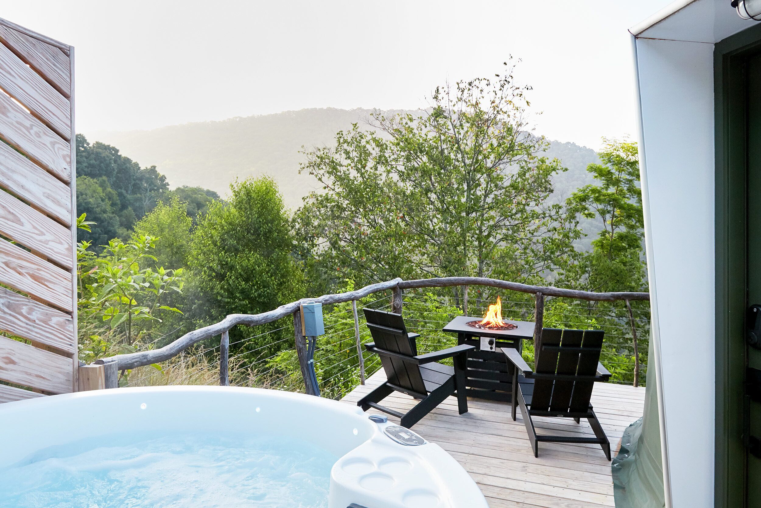 A hot tub on a wooden deck with two chairs and a table, overlooking a forested hillside.