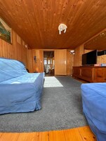 Una habitación con paredes y techo de madera, alfombra gris, dos sofás cubiertos de azul, un mueble de madera con un televisor, y un cuadro en la pared.
