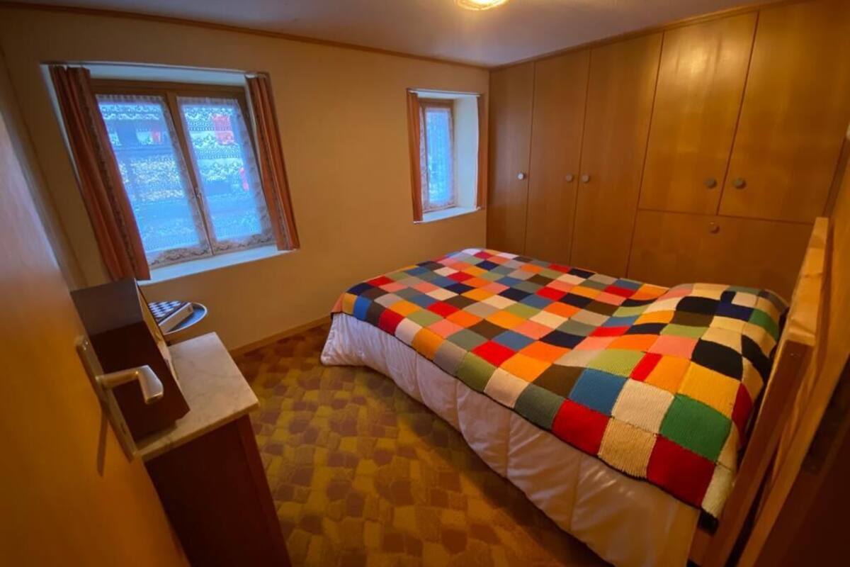 A bedroom with a bed, two windows, a wooden wardrobe, and a small desk with a chair.
