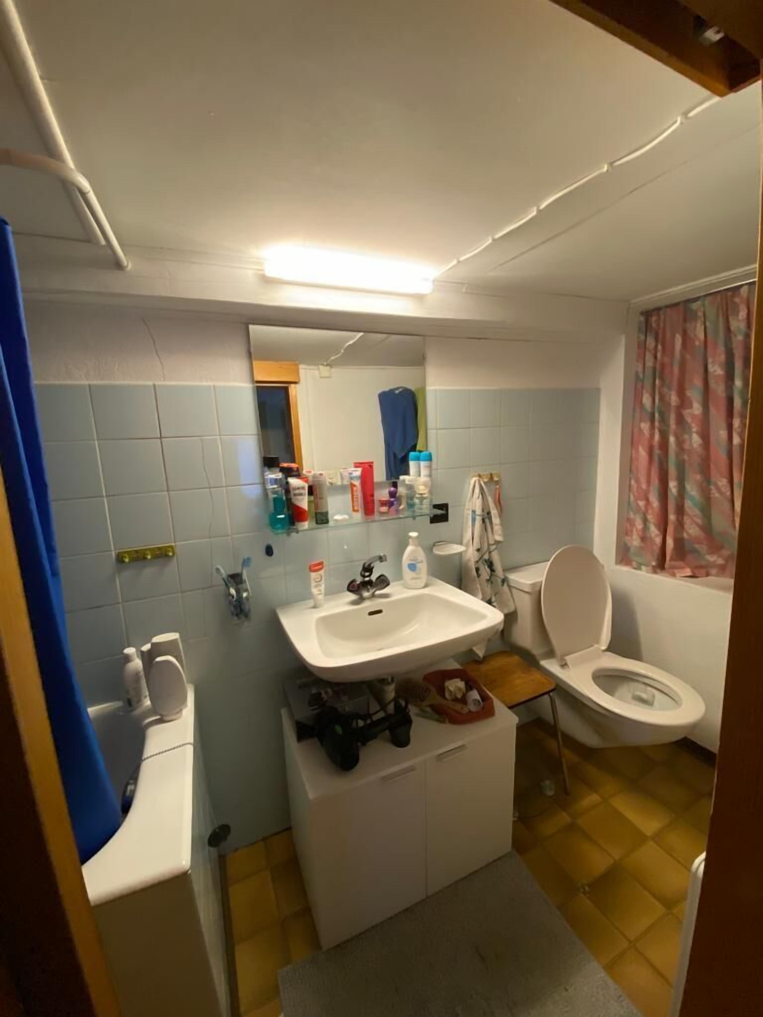 Un baño pequeño con un lavamanos blanco, inodoro y espejo sobre el lavamanos. Hay artículos de tocador en el lavamanos y una toalla colgada en la pared.