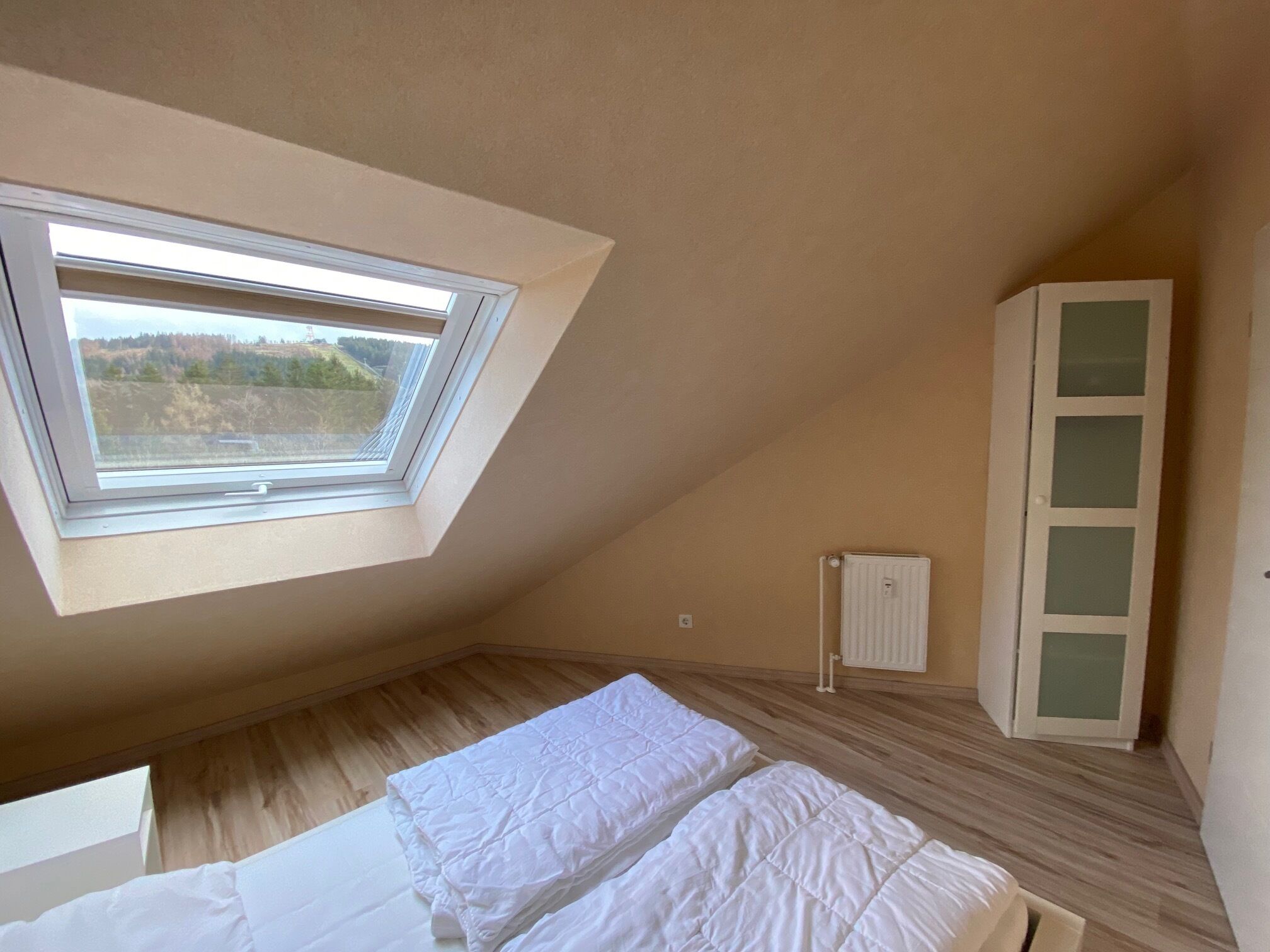 Een slaapkamer met schuine balken, dakraam, bed met witte beddengoed, een wit kastje en een radiator.
