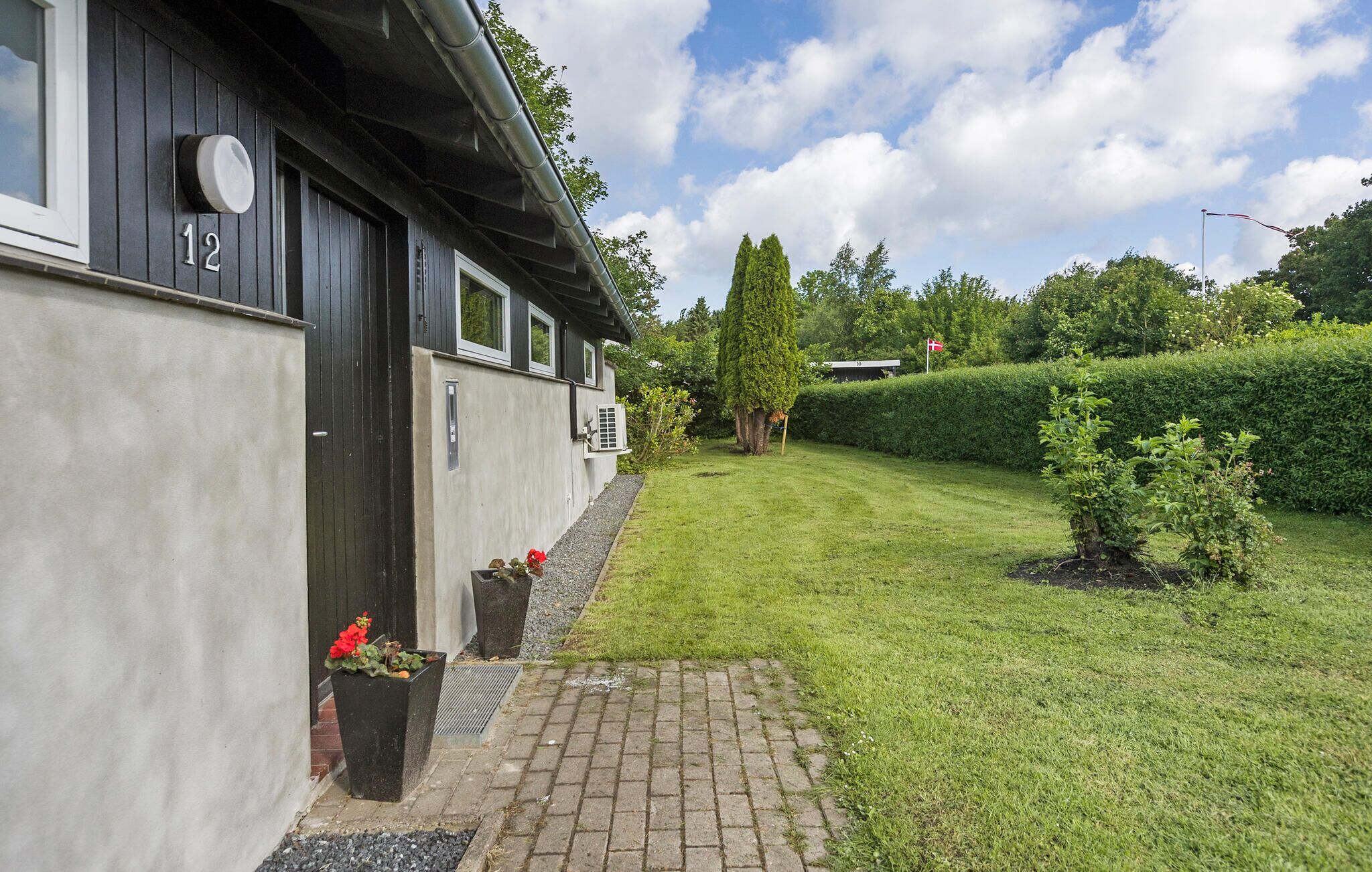 Un bâtiment avec le numéro « 12 » sur le côté, un petit jardin avec une haie et un ciel dégagé.