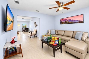 Ein Wohnzimmer mit einem beigen Sofa, einem Couchtisch mit Snacks, einem Fernseher an der Wand und einem Deckenventilator.