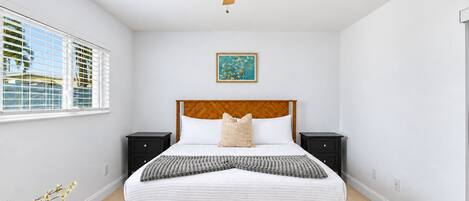 A bedroom with a wooden bed, two bedside tables, a ceiling fan, a window with blinds, and a framed picture on the wall.