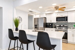 A modern kitchen with white cabinets, stainless steel appliances, a bar area with black chairs, and a ceiling fan.
