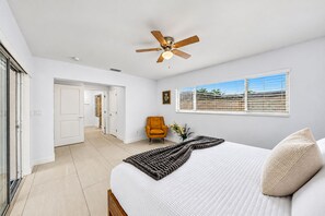 Un dormitorio con cama, ventilador de techo, ventana con persianas y puerta que da a otra habitación.