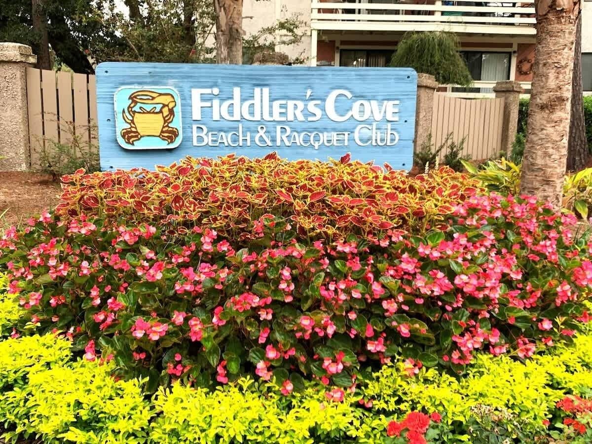 A sign for Fiddler's Cove Beach & Racquet Club with a logo of a crab and a beach scene.