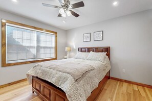 Une chambre à coucher avec un lit en bois, un ventilateur de plafond, une fenêtre avec des stores et une table de chevet avec une lampe.