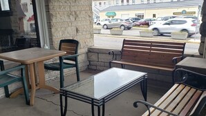 An outdoor seating area with a table and chairs, a bench, and a view of a street with parked cars and a building in the background. - B-town King and Queen (Bloomington)