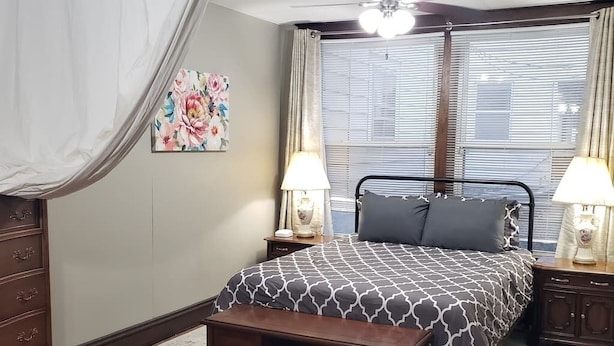 A bedroom with a bed, two bedside tables with lamps, a nightstand, a bench, a window with blinds, and a floral painting on the wall. - B-town King and Queen (Bloomington)