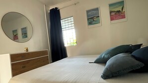 A bedroom with a bed, a dresser, a mirror, a window with curtains, and wall art. - Appartement vue Mer, Rez-de-chaussée Chaussée, Chez Ti Louis - Grande Anse (Les Anses-d'Arlet)