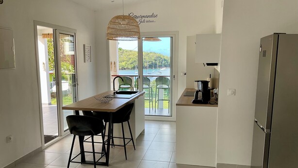 A modern kitchen with a dining area, a view of the sea, and a balcony with green chairs. - Appartement vue Mer, Rez-de-chaussée Chaussée, Chez Ti Louis - Grande Anse (Les Anses-d'Arlet)