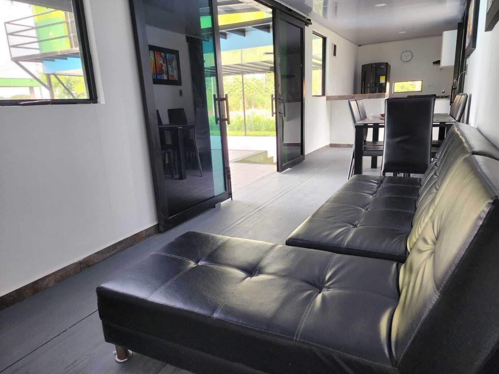 Un interior moderno con un sofá de cuero negro, un ventanal que da al exterior y un comedor con sillas y mesa.