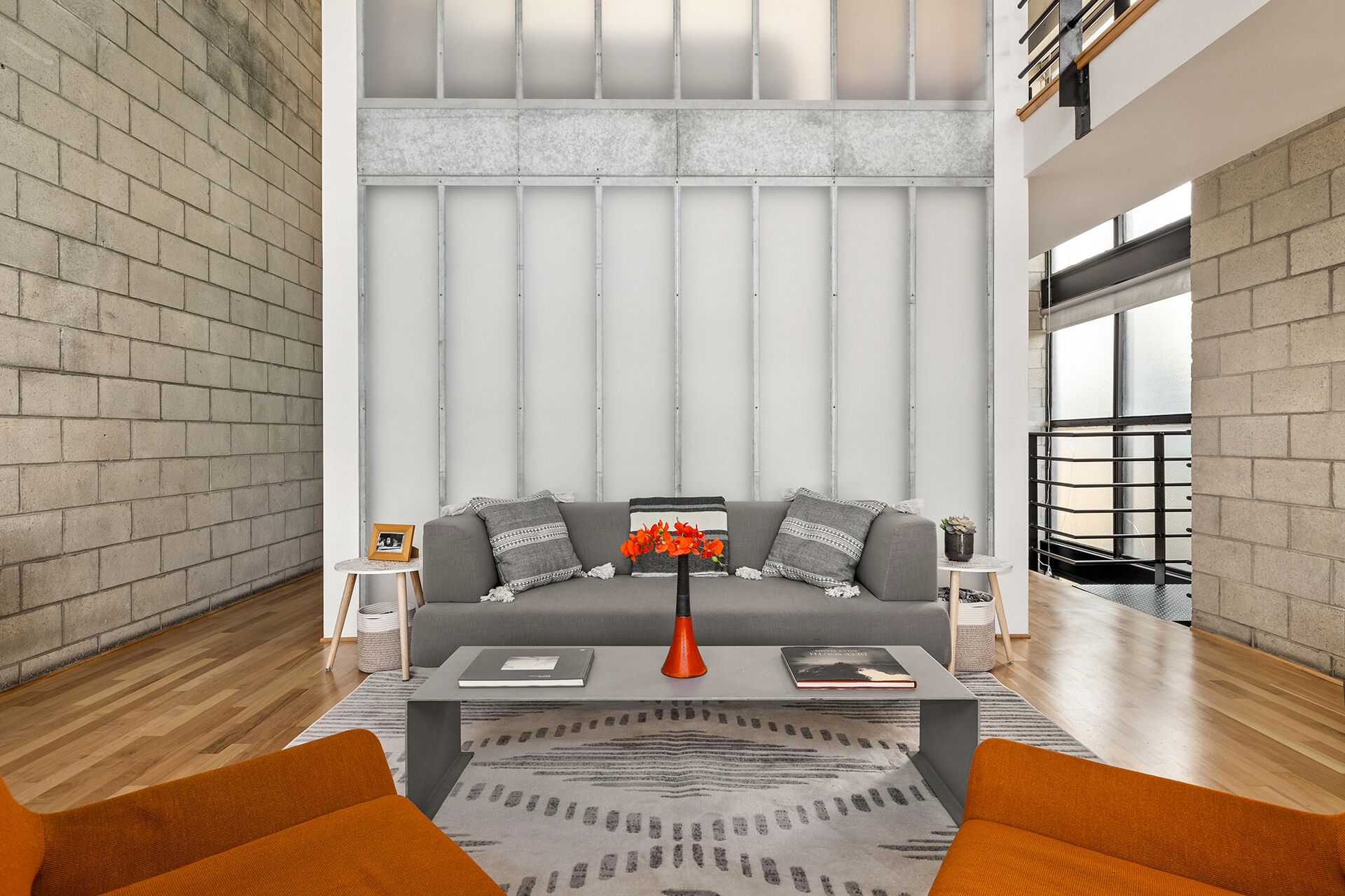 A modern living room with a grey sofa, a coffee table, and orange cushions. There is a small side table with a picture frame and a vase with flowers.
