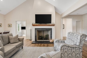 A living room with a fireplace, a sofa, a patterned armchair, and a flat-screen TV mounted above the fireplace.