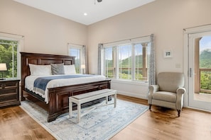 A bedroom with a large bed, a bench, a chair, and a view of the outdoors through large windows.
