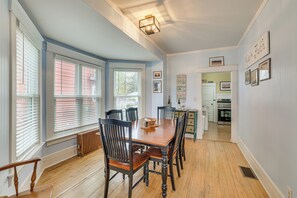 A dining area with a wooden table and chairs, large windows with blinds, a kitchen in the background, and a wall with framed pictures and a sign. - Ludington Home w/ 6 Bicycles < 1 Mi to Beach! (Ludington)