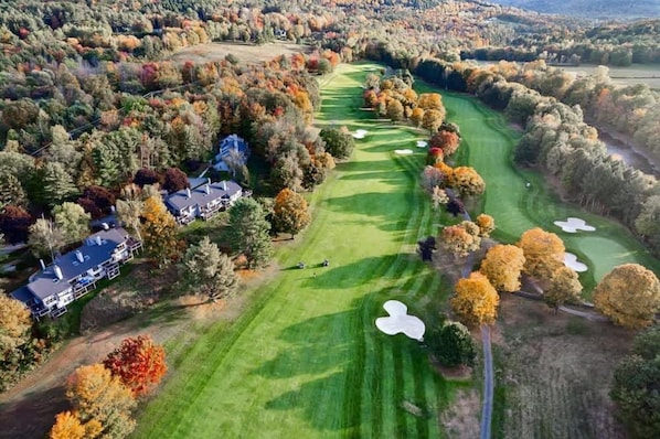An aerial view of a golf course with a clubhouse, surrounded by autumn foliage.
