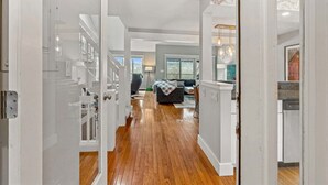 Un couloir doté d’un plancher en bois, de murs blancs et offrant une vue sur un espace de vie comprenant un canapé et une table à manger.