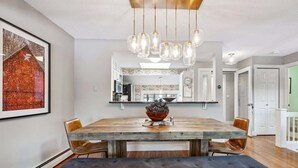 Un coin repas avec une grande table en bois, des chaises, un bol de pommes de pin et un luminaire suspendu.