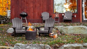 An outdoor seating area with Adirondack chairs, a fire pit, and a grill against a red wooden wall. - Upscale Cabin 1: hot-tub, private firepit, complimentary kayaks + paddleboards! (Cavendish)