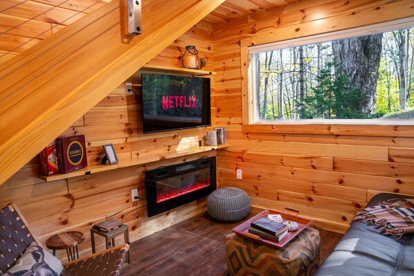 Interior acogedor de una cabaña con una TV mostrando 'NETFLIX', una chimenea, paredes de madera, un ventanal con vistas a un bosque y una zona de estar confortable.