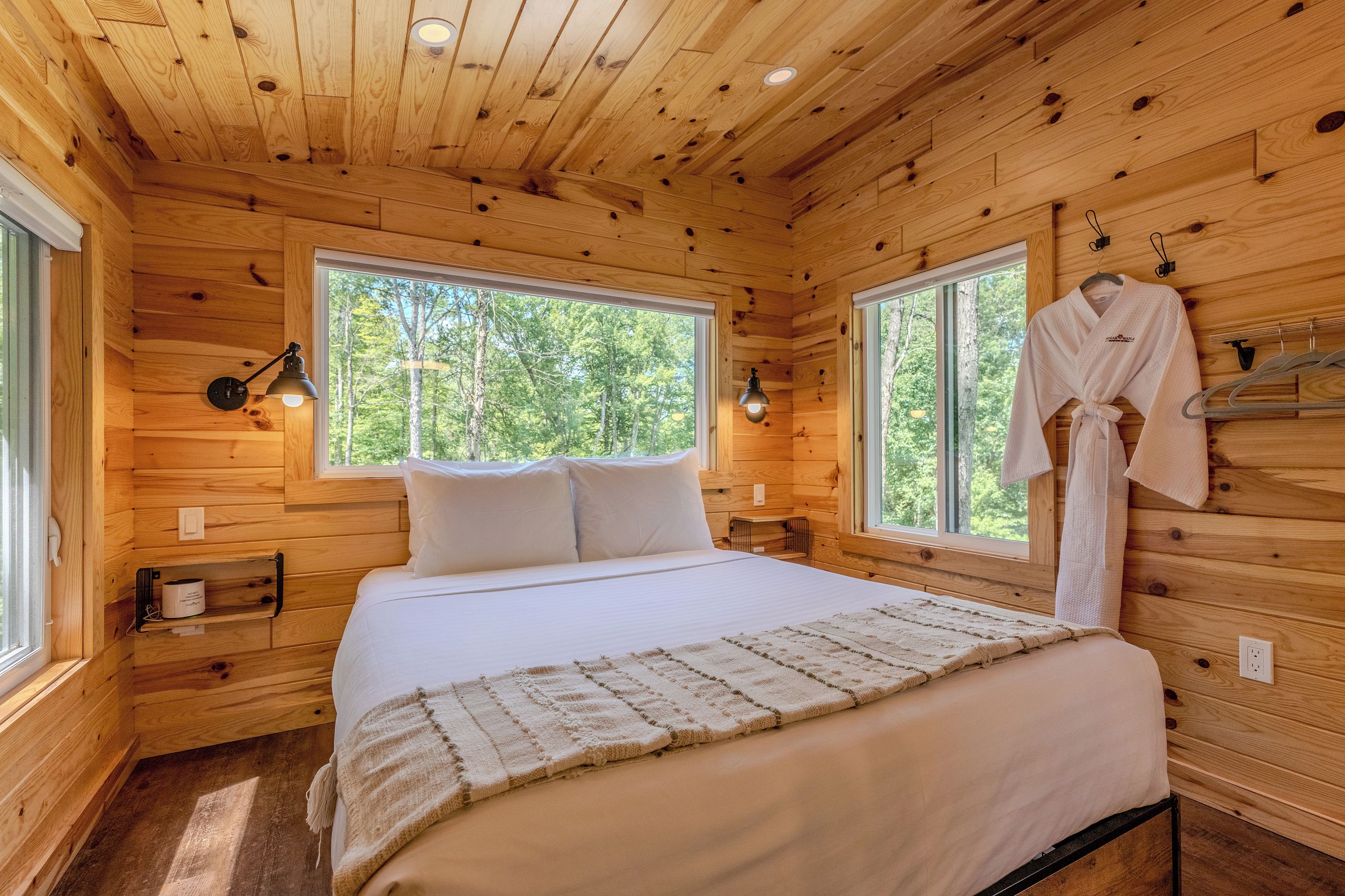 Ein Schlafzimmer in einer Holzhütte mit einem Bett, zwei Nachttischlampen, einem aufhängbaren Bademantel und einem Fenster mit Blick auf Bäume.