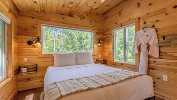Ein Schlafzimmer in einer Holzhütte mit einem Bett, zwei Nachttischlampen, einem aufhängbaren Bademantel und einem Fenster mit Blick auf Bäume.
