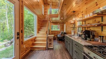 A cabin interior with a wooden staircase, kitchen area, and large windows offering a forest view.