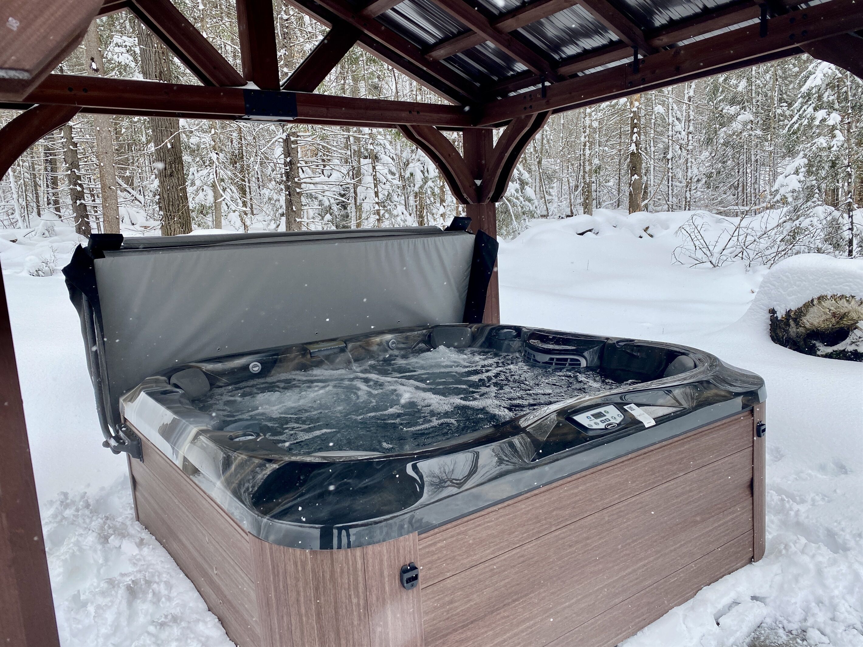 Un bain à remous situé dans un espace extérieur abrité, doté d’une structure en bois et surplombant une forêt enneigée.
