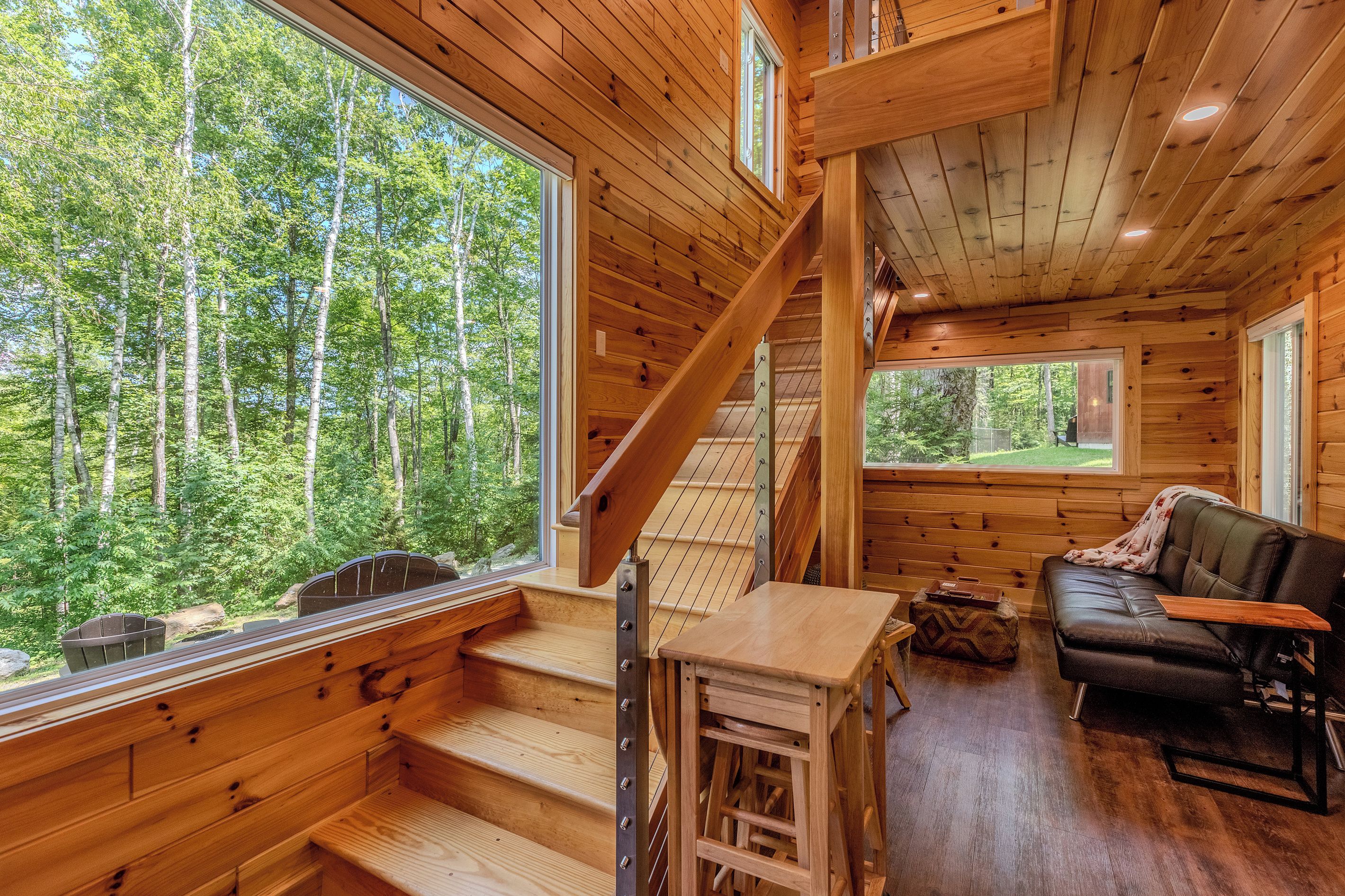 A wooden cabin interior with a staircase, a sofa, and large windows offering a forest view.