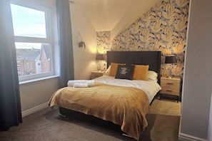 A bedroom with a large bed, a nightstand, a lamp, and a view of buildings through a window. - Avondale, Promenade 2bedroom, free parking & wifi (Southport)
