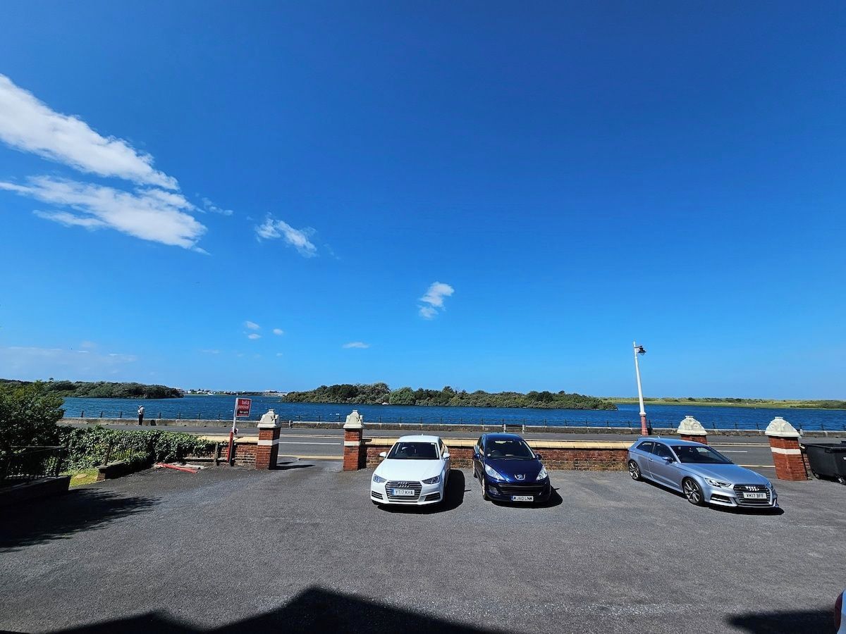 A parking area with three cars, a clear sky, and a body of water in the background.