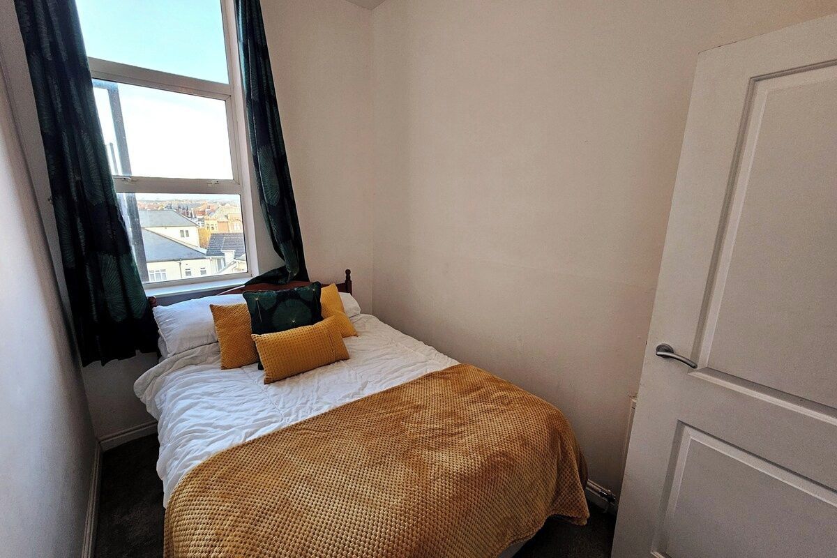 A small, single-bed room with a window, a door, and a view of buildings outside.