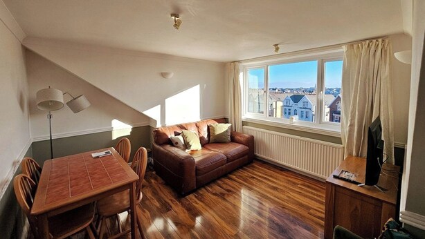Un salón con un sofá de cuero marrón, una mesa de comedor de madera con sillas, un televisor sobre un mueble de madera y una ventana con vistas a edificios.