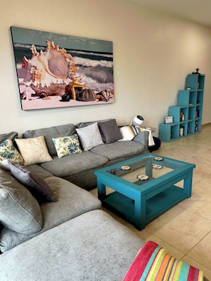 A living room with a grey sectional sofa, a teal coffee table, a colorful striped rug, a blue bookshelf, and a large wall art depicting seashells.