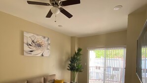 Un salon spacieux doté d’un parquet en bois foncé, d’un canapé beige agrémenté d’un coussin jaune, d’un ventilateur de plafond et d’une grande fenêtre donnant sur un espace clos.