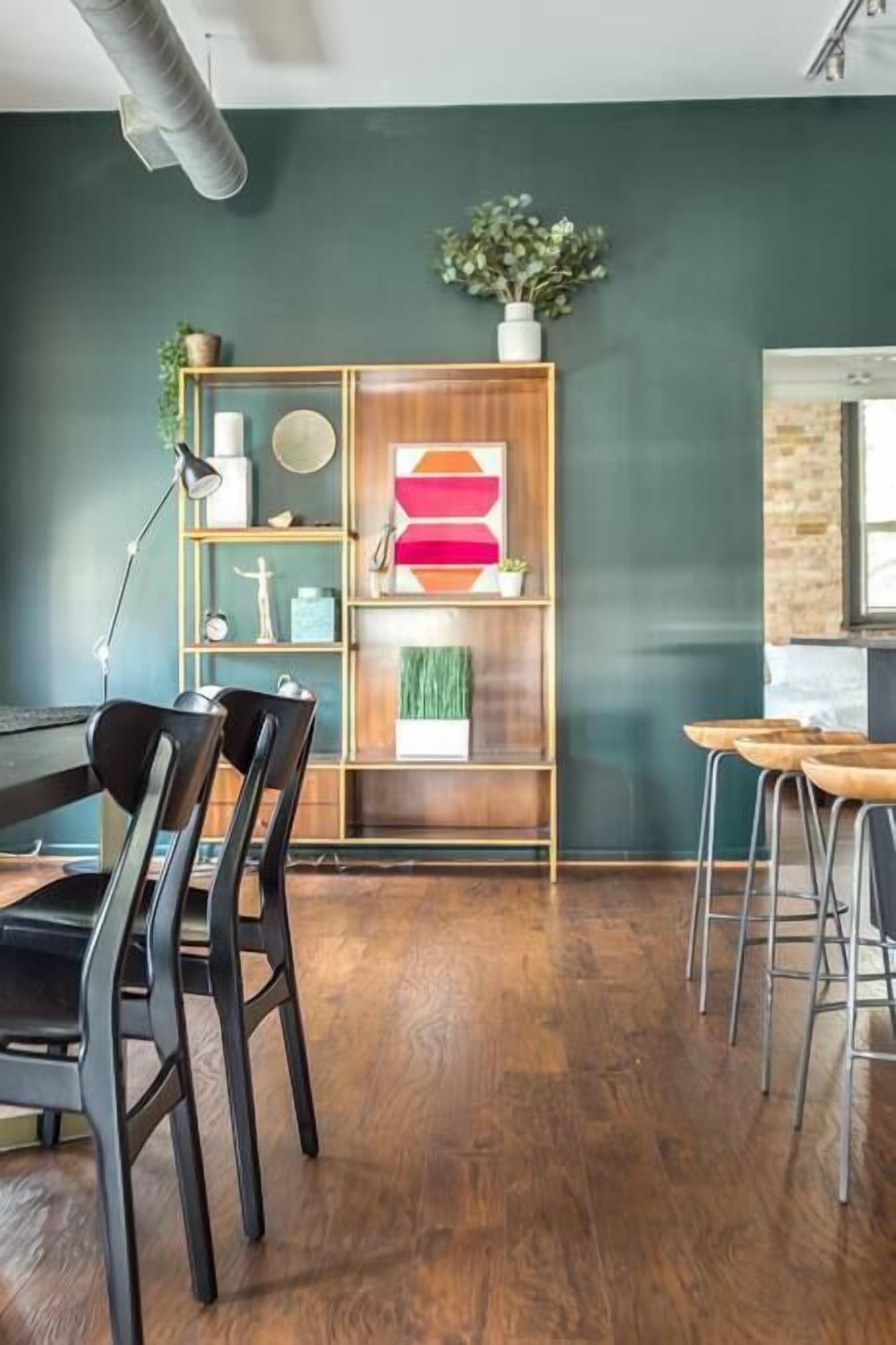 A modern dining area with a wooden table and chairs, a bookshelf with decorative items, and a green wall.