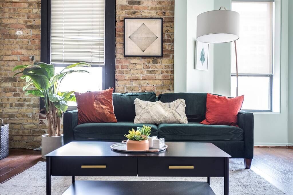 A living room with a dark green sofa, red and white cushions, a coffee table with a plant, and a large window with blinds.
