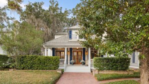 A two-story house with a front porch, surrounded by well-maintained hedges and trees. - Dual Primary Suites & Luxe Whimsical Interiors (Bluffton)